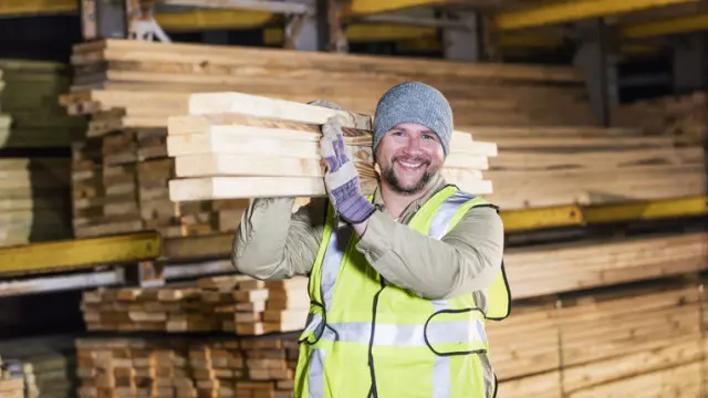 A mid adult Hispanic man in his 30s working at a warehouse at a lumberyard or home improvement store. He is carrying a stack of lumber on his shoulder, smiling at the camera.